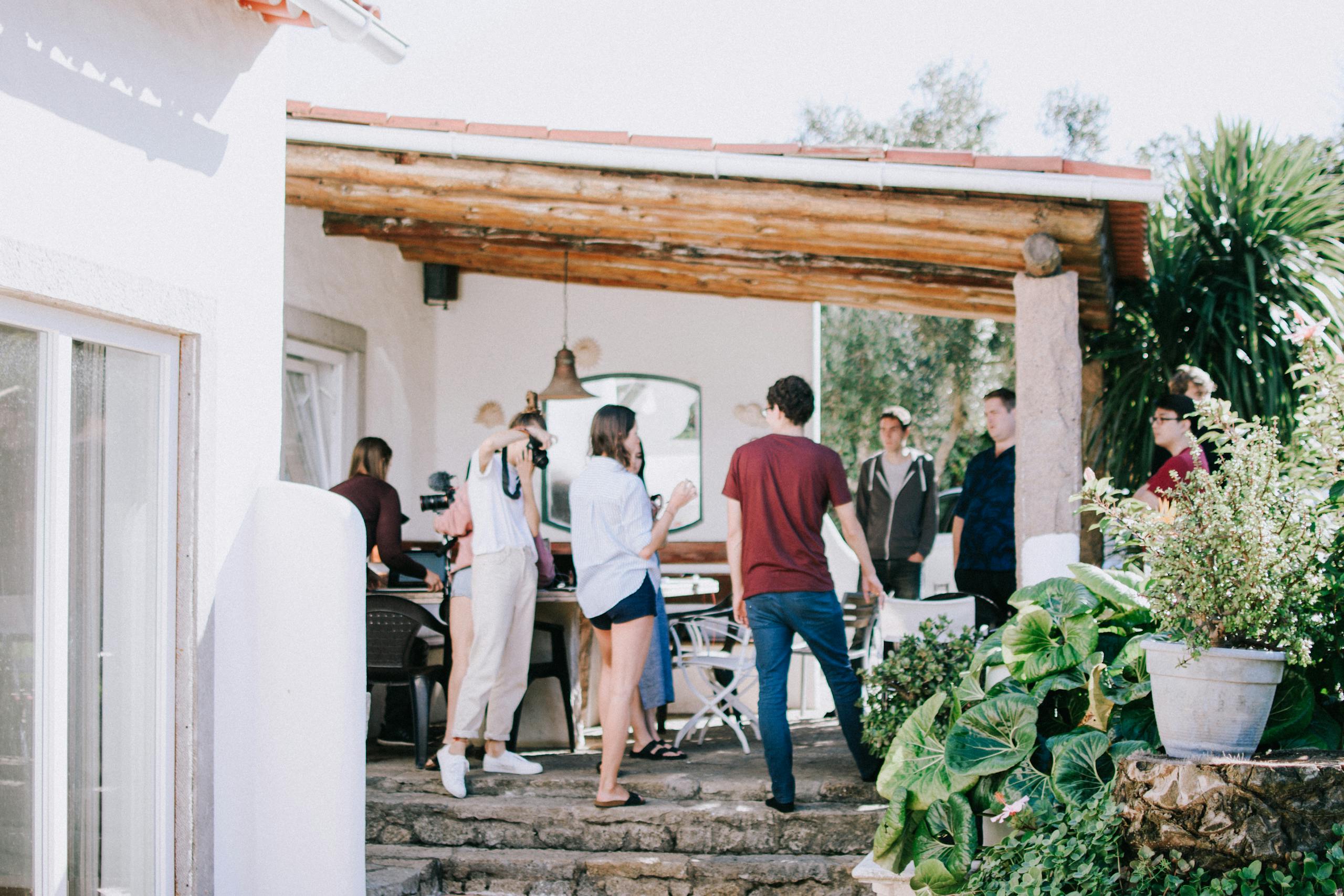 A group of young adults socializing outdoors on a sunny patio, surrounded by greenery.